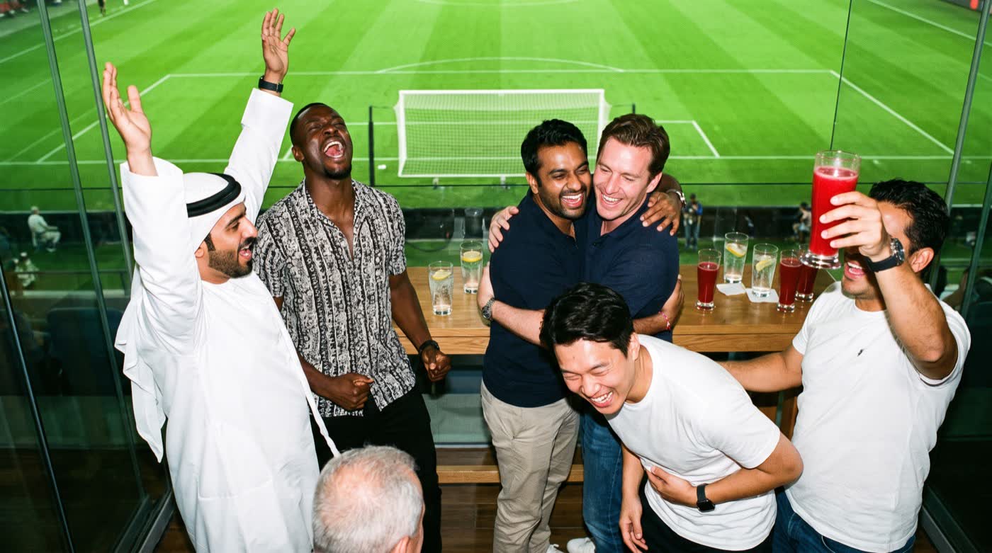 Men in a private box above a stadium celebrating a goal.